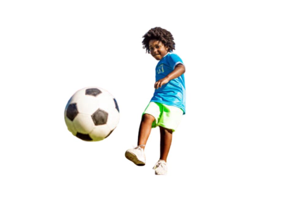 A happy boy in blue jersey and green shorts kicks the soccer ball at us with a grin.